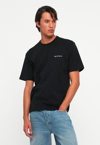 Mann iført svart Belstaff T-skjorte og lyseblå jeans, med den ene hånden i lomma, ser til siden mot en enkel hvit bakgrunn.