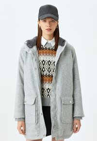 Cappotto in fleece grigio con chiusura a zip e due tasche frontali, indossato sopra un maglione a fantasia con colori arancione, nero e crema. Cappello grigio.