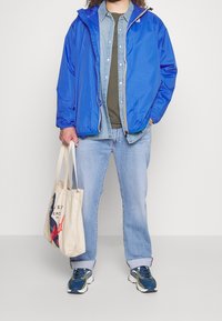 Blauwe lichtgewicht jas met capuchon, groene blouse eronder, licht denim spijkerbroek en marineblauwe sneakers. Beige boodschappentas met grafische print.