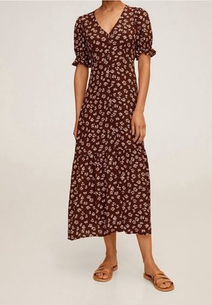 Kvinde iført en brun midi-kjole med hvide blomstertryk, korte puffede ærmer og brune flade sandaler, stående mod en ensfarvet baggrund.