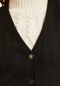 Gilet a righe nere con scollo a V, caratterizzato da un bottone lucido con un motivo a cuore, indossato sopra un maglione a collo alto in maglia cable color crema.
