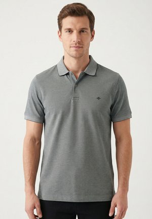 Mann trägt ein graues Polohemd mit kurzen Ärmeln und kleinem Logo auf der Brust, steht vor einem schlichten hellen Hintergrund.