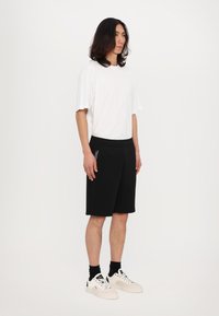 T-shirt bianco a maniche corte abbinato a pantaloni corti neri con vita regolabile e tasche laterali, indossato con scarpe da ginnastica bianche e calze nere.