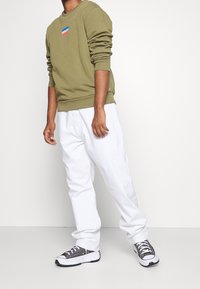 Sweat-shirt vert olive avec poignets et encolure côtelés, arborant un petit logo Levi's multicolore ; porté avec un pantalon blanc ample et des baskets grises.