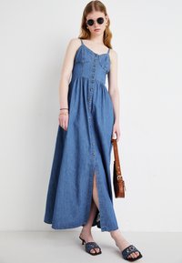 Denim maxi kjole med en tettsittende overdel, tynne stropper og knappelukking foran. Har en høy splitt til låret og en flytende skjørt, i en lys blå farge.
