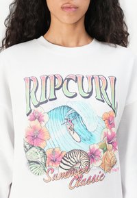 Felpa bianca con un grafico colorato che presenta il testo "RIPCURL", un'onda, un surfista, fiori e conchiglie. La texture è liscia.