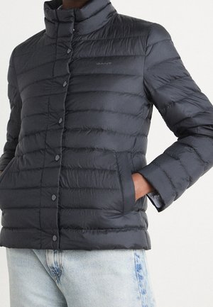 Personne portant une veste matelassée noire GANT avec boutons-pression et poches latérales, associée à un jean en denim bleu clair.