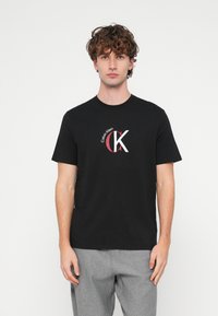 Czarny bawełniany t-shirt z okrągłym logo Calvin Klein w kolorze białym i czerwonym, z okrągłym dekoltem i krótkimi rękawami, noszony z szarymi spodniami.