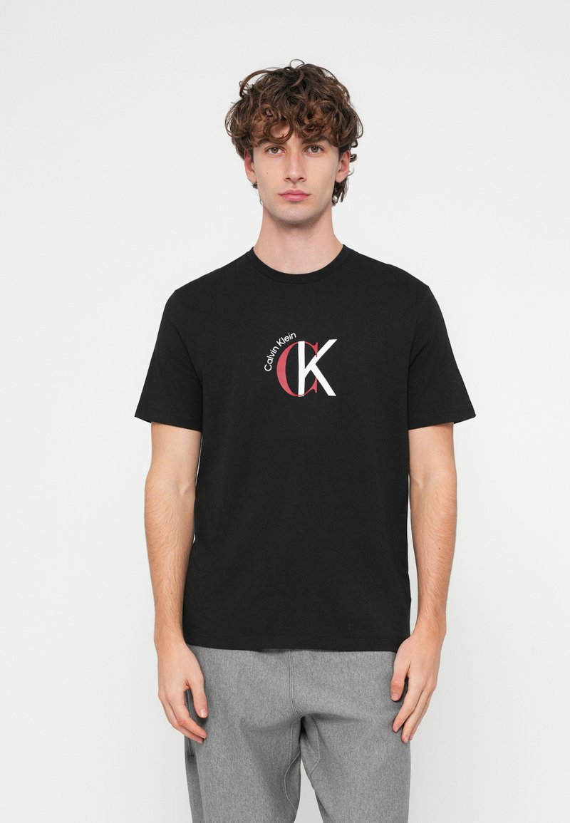 Czarny bawełniany t-shirt z okrągłym logo Calvin Klein w kolorze białym i czerwonym, z okrągłym dekoltem i krótkimi rękawami, noszony z szarymi spodniami.