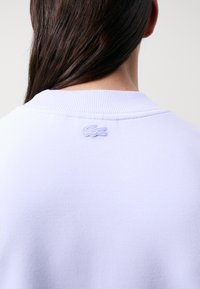 Gaiši purpura krekls piedāvā ribotu apkakli, teksturētu audumu un smalku izšūtu logo aizmugurējā apkakles daļā.