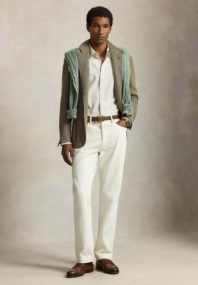 Homme portant un pantalon blanc, une chemise beige, un blazer marron texturé, un pull vert drapé sur les épaules, une ceinture marron, une montre et des mocassins marron, debout.