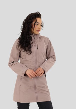 Pitkäkiharainen nainen, jolla on vaalean mauve väriin vivahtava korkeakauluksinen vetoketjullinen takki, kädet yhteenliitettyinä, katsoo sivulle harmaata taustaa vasten.