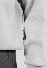 Sudadera gris con una textura suave, puños acanalados y una etiqueta negra con "PROHIBIDO" en la costura lateral. Diseño casual con mangas largas.