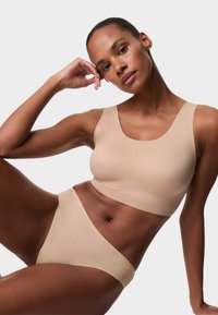 Mujer recostada con un brazo apoyado en la cabeza, vestida con un sujetador beige sin costuras y ropa interior a juego sobre un fondo claro y liso.