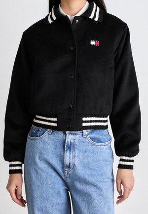 Vrouw draagt een zwart varsity-stijl jasje met witte gestreepte kraag, manchetten en zoom, gecombineerd met lichtblauwe, hoog getailleerde jeans.