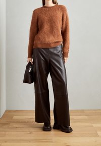 Brązowy puszysty sweter z okrągłym dekoltem, w połączeniu z szerokimi spodniami skórzanymi w kolorze czarnym. Modelka trzyma małą czarną torebkę, stojąc na drewnianej podłodze.
