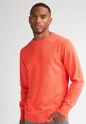 Man wearing a bright orange crewneck sweater with ribbed cuffs and hem, standing against a plain light background.