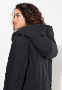 Svart vadderad jacka med huva, med ett quiltat mönster och synliga sömmar. Inkluderar en märkespatch på ärmen.