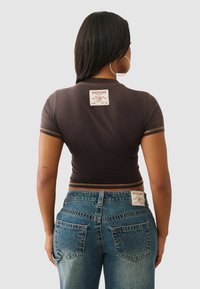 Brązowy krótki t-shirt z krótkimi rękawami, złotą nicią i naszywką z logo; w połączeniu z niebieskimi dżinsami z charakterystycznym wzorem na tylnej kieszeni.