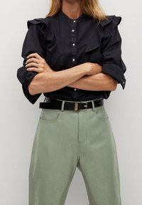 Blusa negra con volantes, cierre de botones y mangas enrolladas, combinada con pantalones de tiro alto verdes claros y un cinturón negro con accesorios dorados.