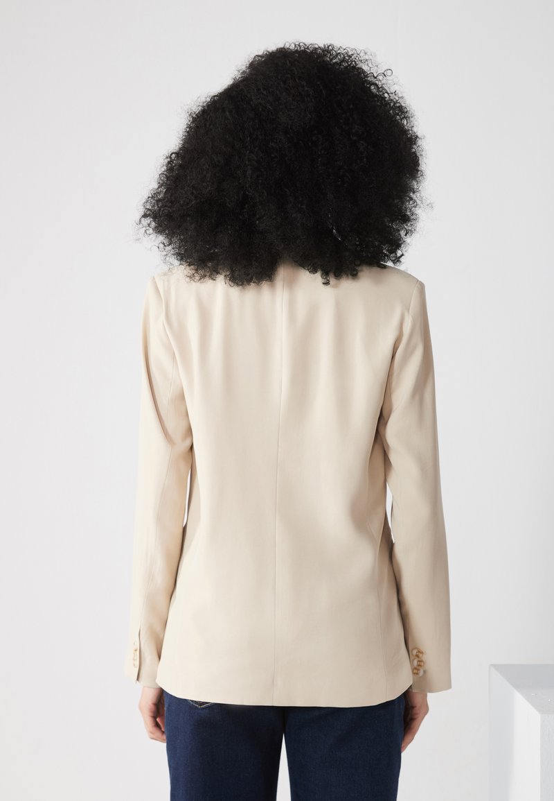 Blazer beige su misura con maniche lunghe, spalline sottili e bottoni sui polsini, indossato sopra pantaloni in denim scuri.