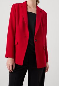 Červený blazer vyrobený z hladké tkaniny s strukturovaným střihem. Obsahuje ostré klopy, uzávěr na jeden knoflík a kapsy vpředu.