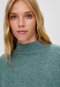 Zielony sweter o teksturowej powierzchni z wysokim dekoltem. Miękka tkanina z nakrapianym wzorem. Posiada dopasowany kołnierz i luźny wygląd.