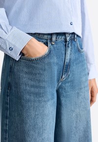 Osoba w niebieskiej, prążkowanej koszuli i niebieskich dżinsach z lewą ręką w kieszeni na białym tle.