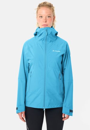 Donna che indossa una giacca impermeabile Vaude azzurro chiaro con tasche con cerniera, colletto alto e pantaloni neri, in piedi su sfondo bianco.