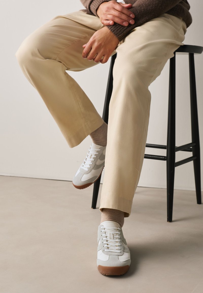 Personne assise sur un tabouret noir portant un pantalon beige, des chaussettes marron, des baskets blanches et grises, et un pull marron avec les mains reposant sur le genou.