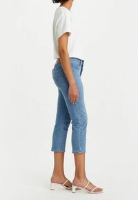 Jeans de mezclilla azul claro con un ajuste recortado, que presentan bolsillos traseros clásicos, detalles de costura y una cintura de altura media, combinados con sandalias blancas de tipo slide.