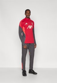Rød og grå atletisk træningsdragt med høj krave, der har et AXA-logo. Sorte sportssko fuldender outfittet.