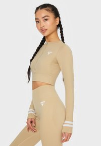 Beige sportlicher Crop-Top mit Reißverschluss, langen Ärmeln und Logo; kombiniert mit hoch taillierten Leggings mit weißen Streifen.
