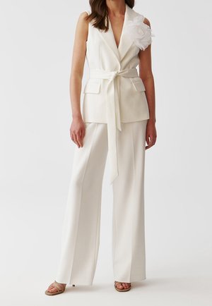 Woman wearing sleeveless white belted blazer with large fabric flower on shoulder and matching wide-leg trousers with open-toe heels.