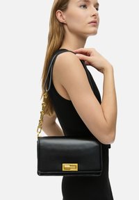 Černá kožená kabelka se zlatým řetízkovým popruhem a obdélníkovou sponou. Hladký povrch, minimalistický design a strukturovaný tvar.