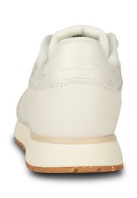 Zadní pohled na bílé tenisky s texturovanými bočními panely, polstrovaným límcem, béžovou mezipodešví a hnědou gumovou podešví. Na patě je embosovaný nápis "WODEN".