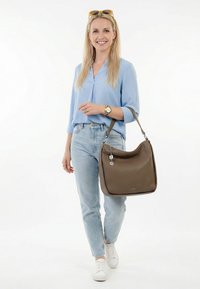Smiling woman in light blue blouse and jeans walking, wearing white sneakers, sunglasses on head, holding large brown leather handbag on arm.