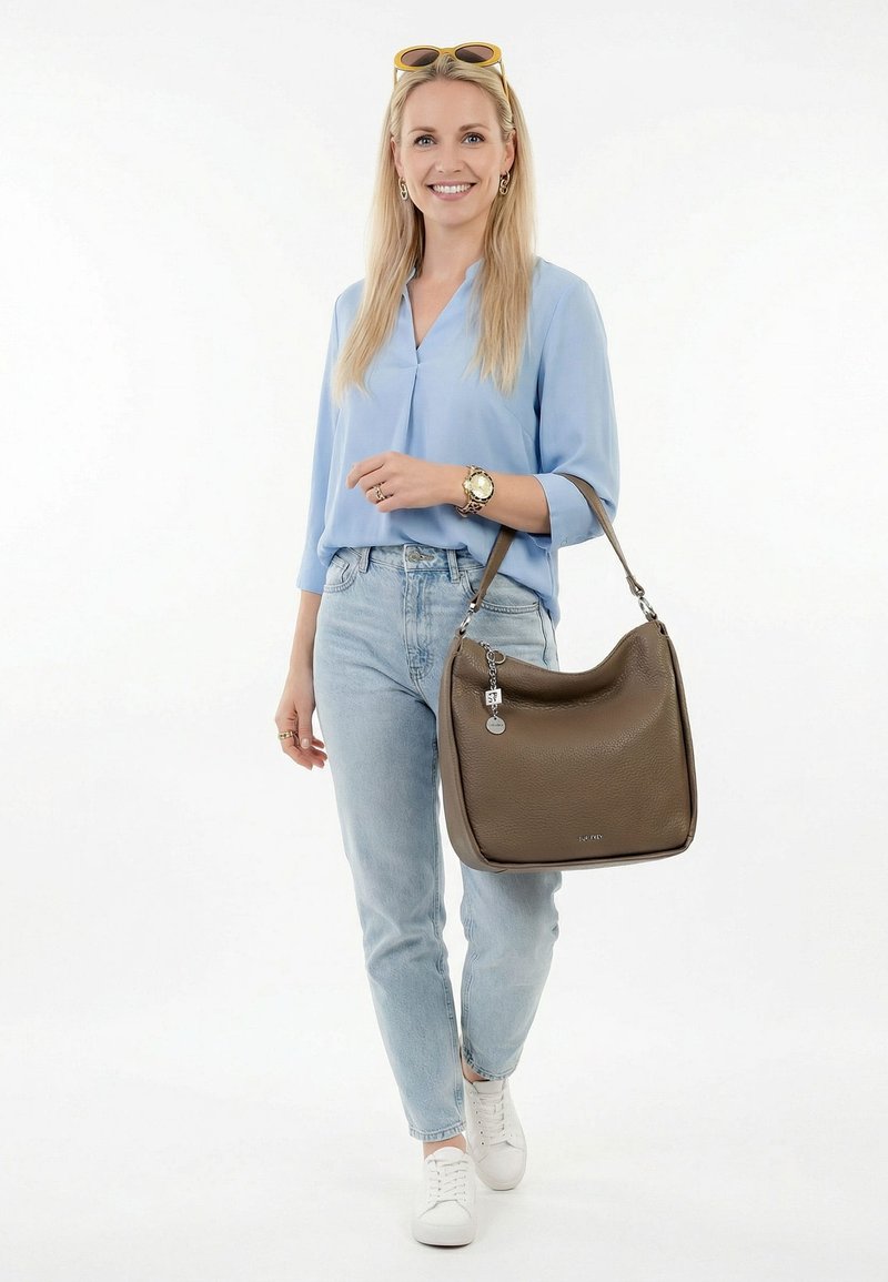 Smiling woman in light blue blouse and jeans walking, wearing white sneakers, sunglasses on head, holding large brown leather handbag on arm.