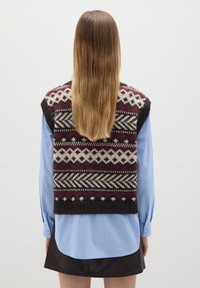 Strickpullover-Weste mit geometrischen Mustern in Bordeauxrot, Creme und Schwarz. Über einem hellblauen Langarmshirt getragen. Textur aus Wollmischung.