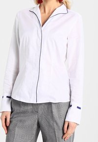 Camisa blanca con cuello de pico, mangas largas y ribete azul marino en los bordes; combinada con pantalones gris con estampado.