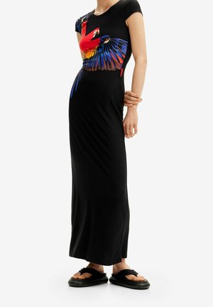 Kobieta ubrana w czarną maxi sukienkę z kolorową grafiką papugi na górnej części przodu, w połączeniu z czarnymi sandałami na platformie.