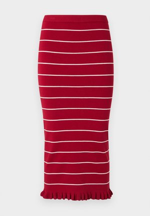 Žena stojaca s prekríženými rukami, oblečená v červenom midi šatách bez rukávov s bielymi pruhmi a čiernych sandáloch na jednoliatom sivom pozadí.