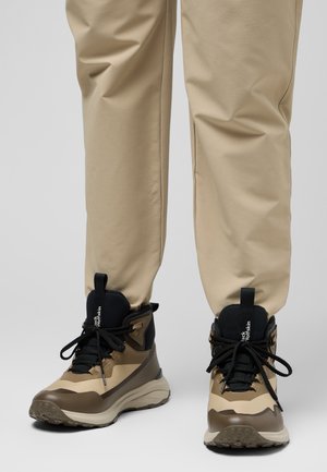 Bottes de randonnée en mélange de matériaux synthétiques beige et marron foncé, avec des lacets noirs et une semelle texturée pour une meilleure adhérence.