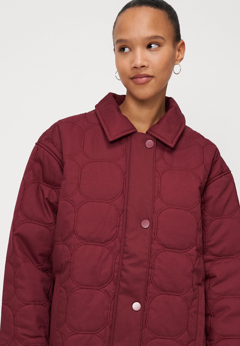 Veste matelassée bourgogne avec des coutures circulaires, texture rembourrée et col. Équipée de boutons-pression et d'une coupe décontractée.