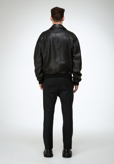 Hombre de espaldas vestido con una chaqueta de cuero negra, pantalones negros y zapatos negros gruesos, sobre un fondo liso.