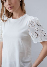 White cotton t-shirt with embroidered eyelet lace sleeves, featuring circular patterns and scalloped edges. Smooth texture, fitted design.