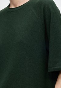 Personne portant un pull en maille vert foncé à manches courtes avec un col rond, montrée de l'épaule jusqu'au milieu du torse.