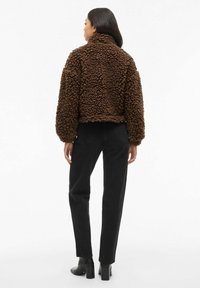 Smeđi, teksturirani, kovrčavi lažni krzneni kaput s visokim ovratnikom, skraćenog kroja i dugim rukavima. Uparen s crnim hlačama visokog struka i potpeticama.