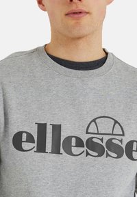 Grauer Sweatshirt mit schwarzem "ellesse"-Logo auf der Brust und geripptem Halsausschnitt. Über einem dunklen Shirt getragen, aus einer Baumwollmischung.