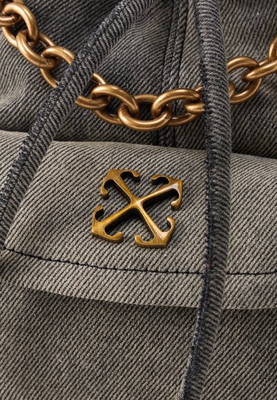 Κοντινή λήψη γκρι υφάσματος με υφή και μαύρο περίγραμμα, με χρυσό εμβληματικό μοτίβο αγκυρών που διαπλέκονται και χοντρή χρυσή αλυσίδα.